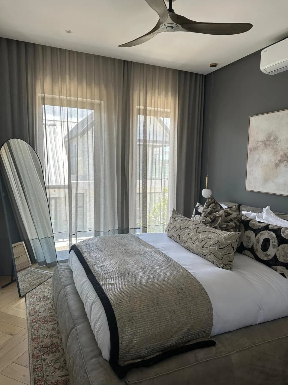 Paarl bedroom with full-height sheer curtains, a floor mirror, layered neutral bedding and a sculptural ceiling fan