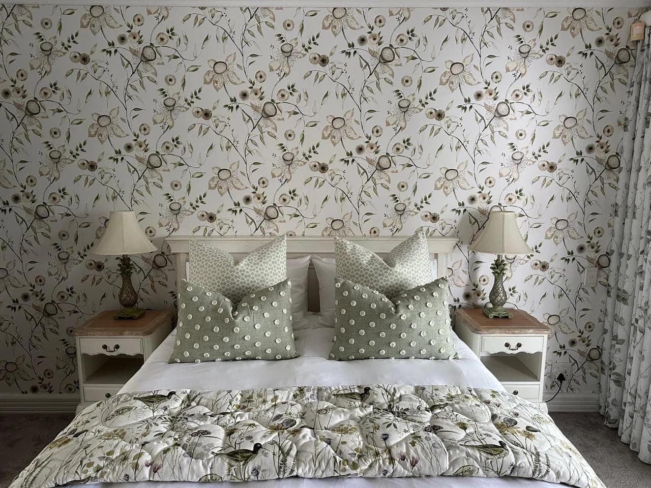 Floral wallpaper in a coordinated bedroom with green scatter cushions, botanical bedding, matching curtains and bedside lamps.