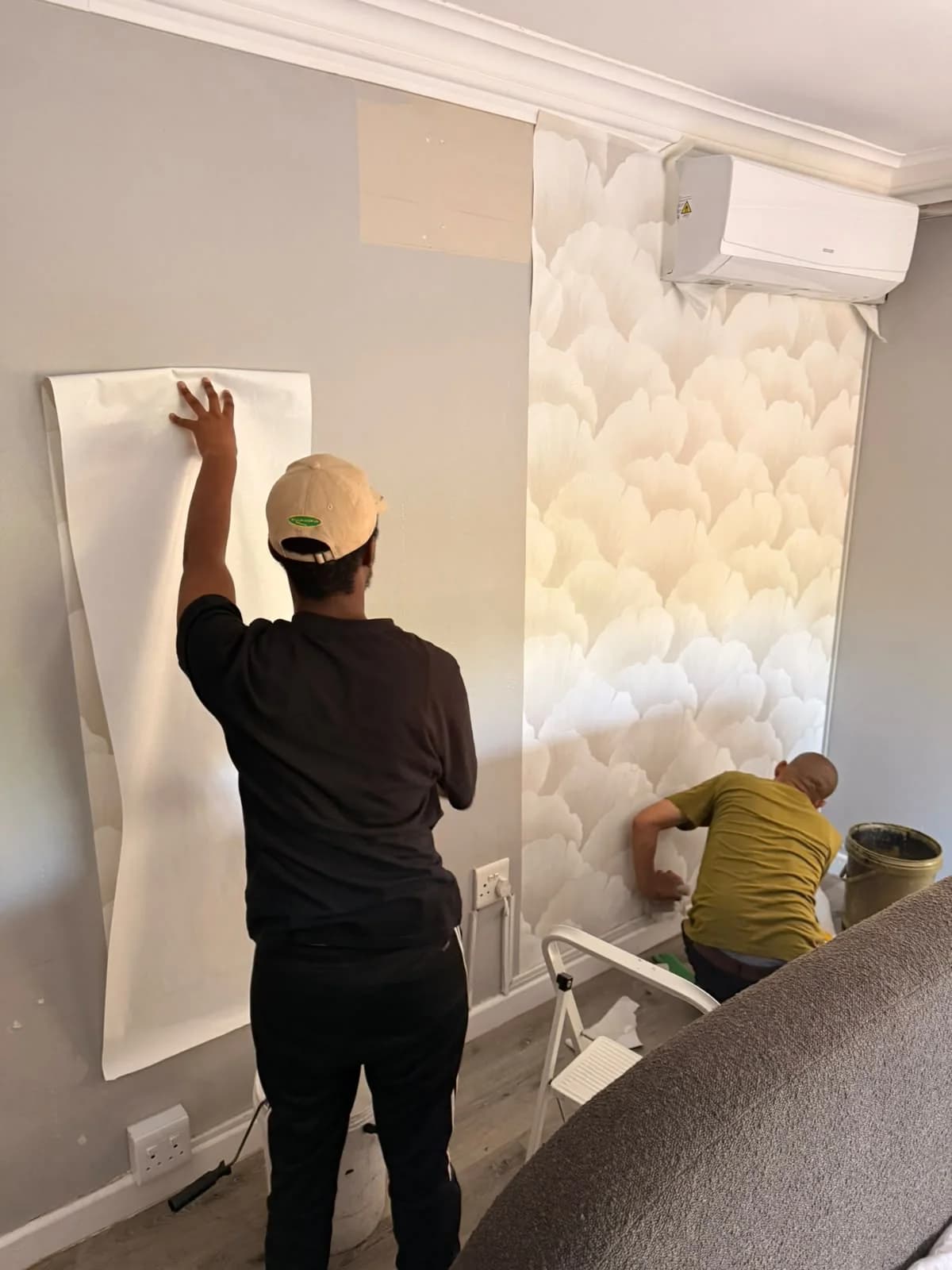 Two installers fitting pale floral wallpaper around a wall-mounted air conditioner.